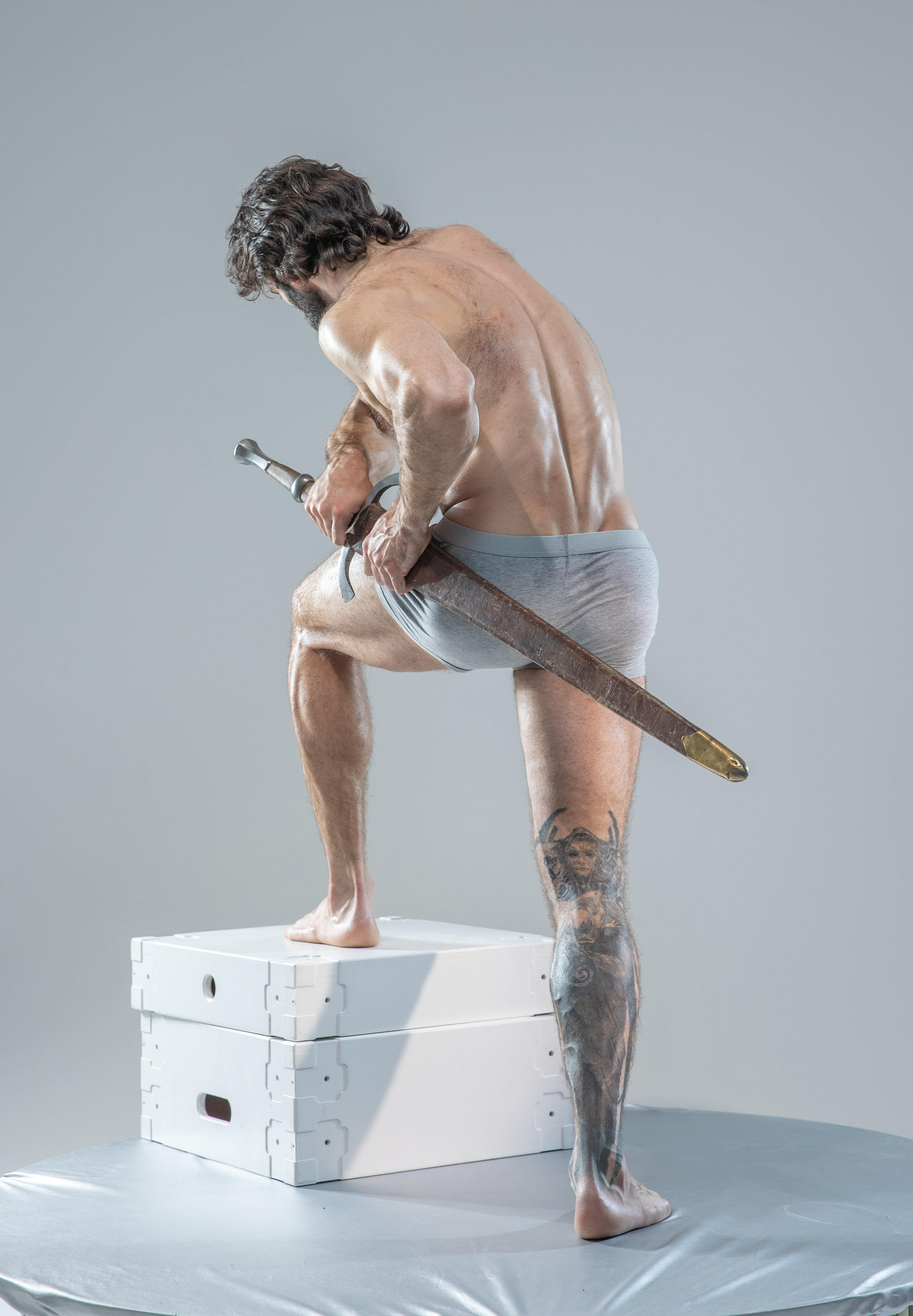 a man with long hair and beard is standing on a white cube with his back to the camera. he is holding a sword in his right hand and is wearing only underwear. the background is a light gray.