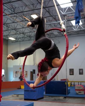 a woman performs aerial hoop, she hangs upside down in the red hoop attached to white rope, she's wearing a black sports bra and black pants and a red fabric is hanging from the ceiling near her. the gym's floor is covered with blue mats, the gym is empty and well lit.