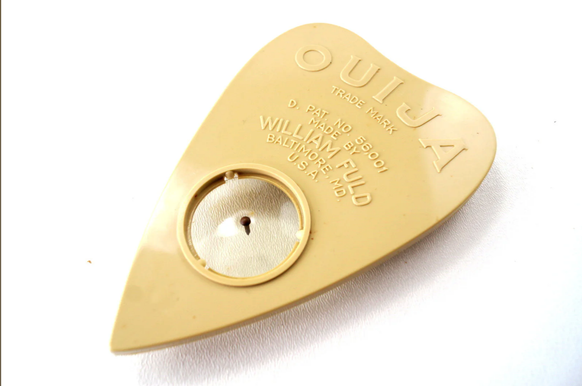 a closeup shot of a classic ouija board planchette, made of a tan plastic, with the trademark "ouija" and manufacturer's information embossed in the plastic. the planchette is designed in the shape of a pointed heart with a round window in the center to hold a planchette. the planchette is sitting on a white background.