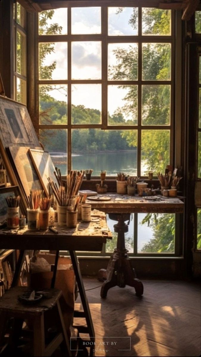 the image showcases an artist's studio filled with natural light. there's a window overlooking a lake surrounded by trees. the studio contains an easel with paintings, a desk filled with various art supplies such as paintbrushes and pots, a stool and the sunlight streams in, creating shadows on the wooden floor.
