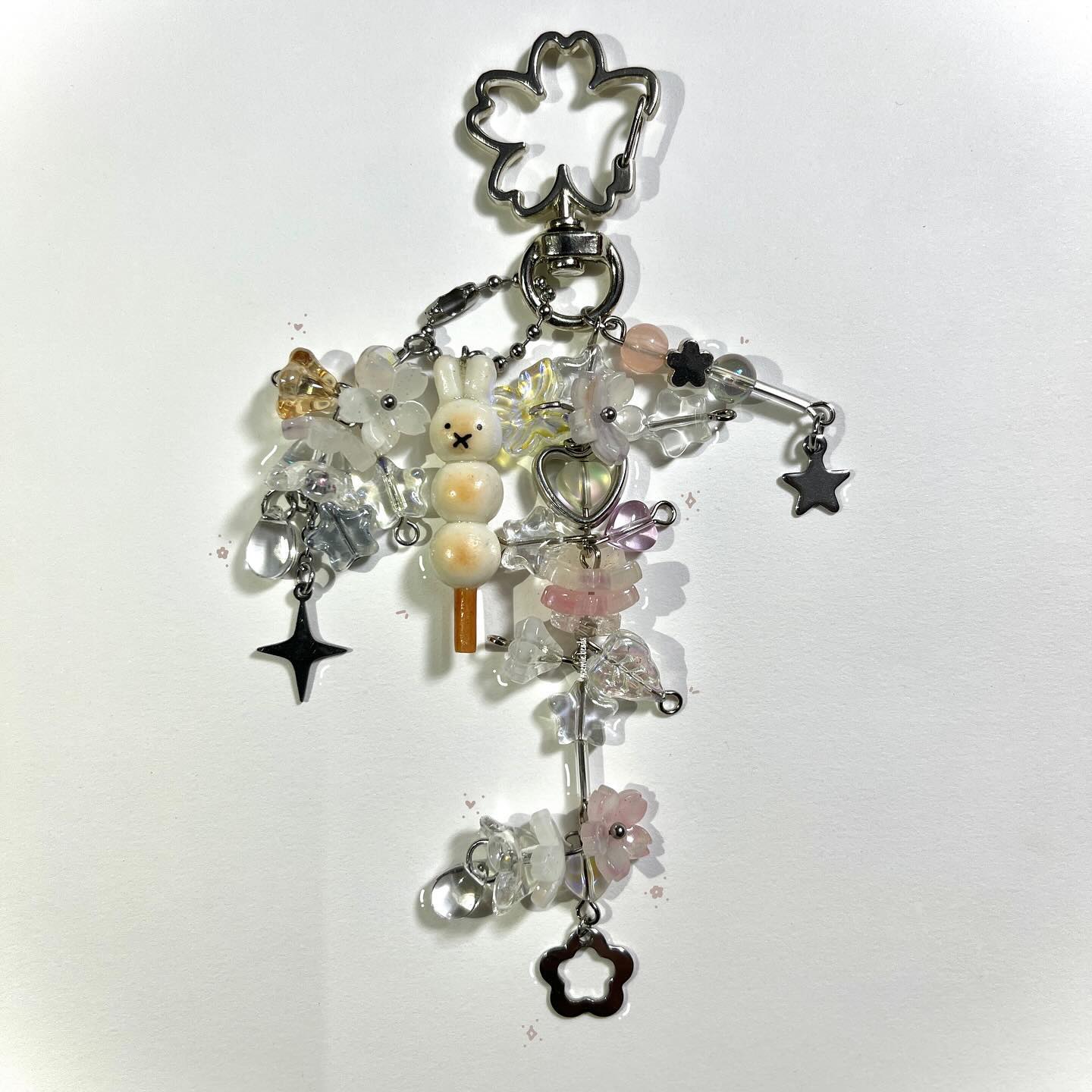 a closeup shot shows a beaded charm or keychain against a white background. the charm is made of various beads and charms attached to silver hardware, including a flowershaped clasp at the top. the charm features a rabbit made of stacked dangoshaped beads with a face on the top bead, as well as flower beads in white and pink shades. other charms include star and heart shapes, and transparent beads. the arrangement creates a playful and whimsical aesthetic.