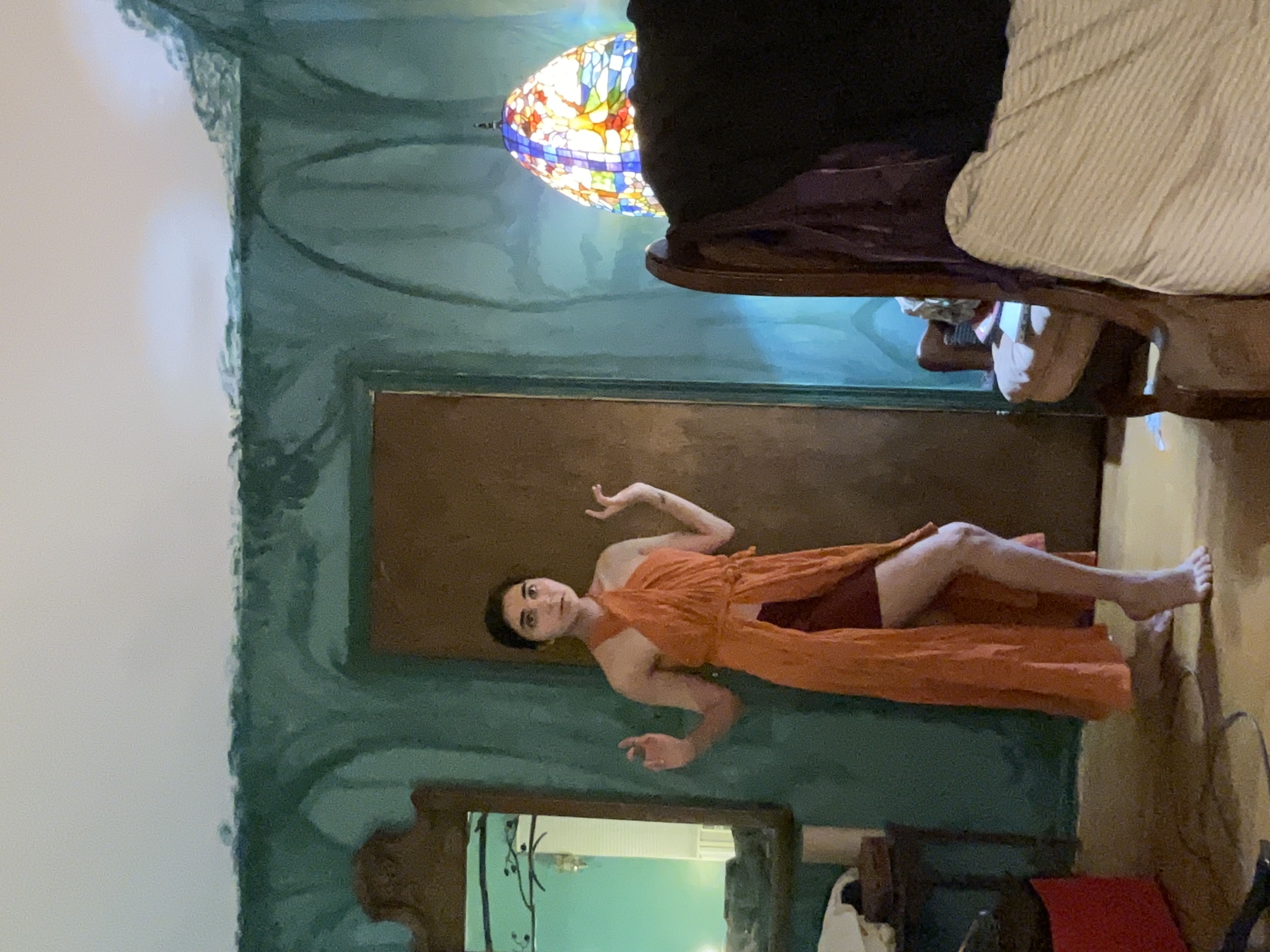 a young woman with dark hair is standing in a room with teal painted walls. she is wearing a long, orange wrap dress with a high slit that shows her legs.  the dress is loosely fitted and appears to be made of a lightweight fabric.  she is striking a pose, with her hands slightly raised.  the background includes vintage furniture, such as a dark wooden bed frame and a vanity with a mirror. an antique stainedglass lamp is visible on a side table. the walls have a mottled, textured appearance, suggesting an artistic paint job. the overall mood is somewhat bohemian.
