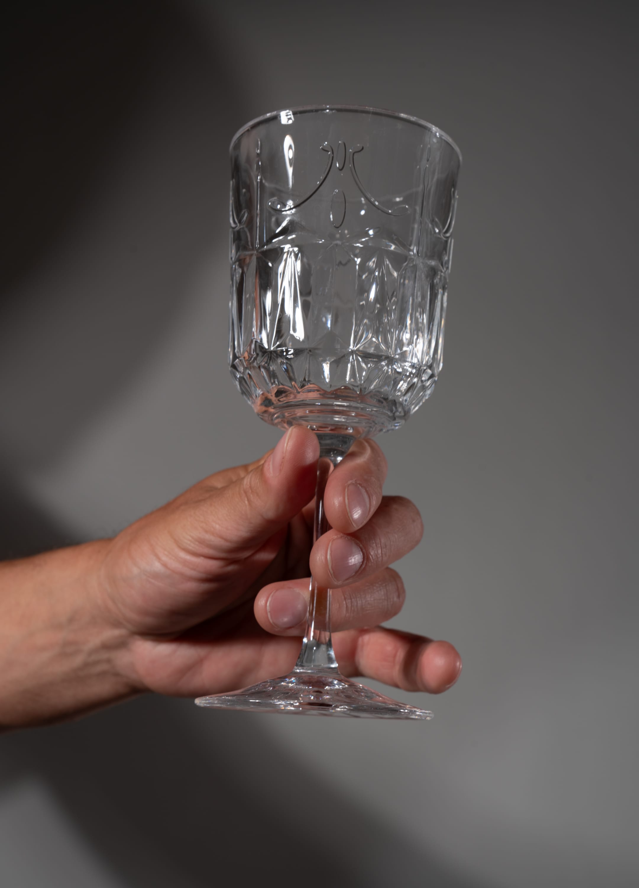 a hand holding an empty clear wine glass against a neutral gray background. the image is in low angle. the glass has a geometric patterned design.