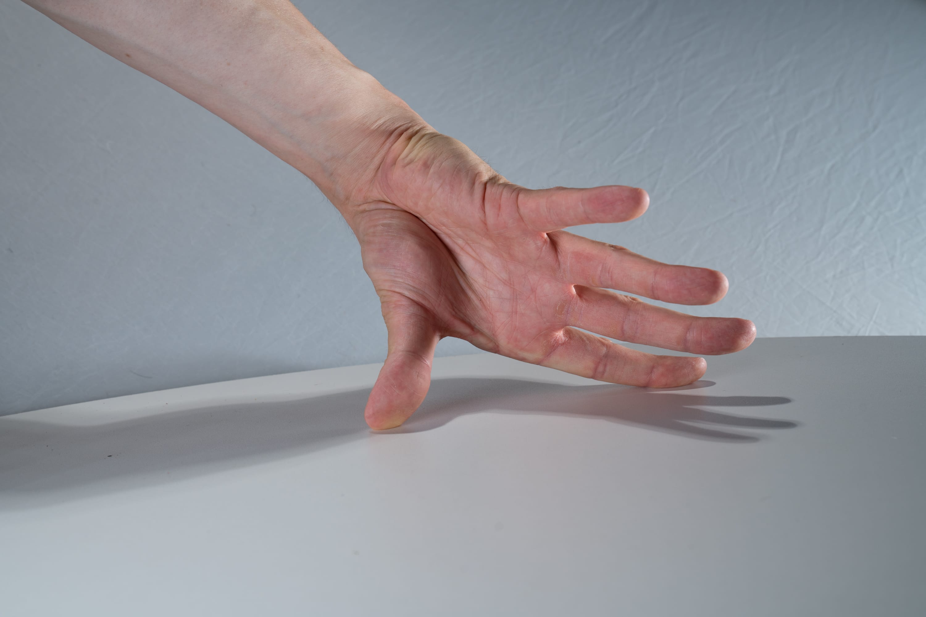 a closeup image of a person's hand and arm extended over a white table. the hand is open, with fingers slightly spread, and the palm facing down. the arm is positioned diagonally across the image, with the hand touching the table near the bottom edge. the table is brightly lit by artificial light, casting a shadow of the hand on the surface. the background is a plain light gray wall.