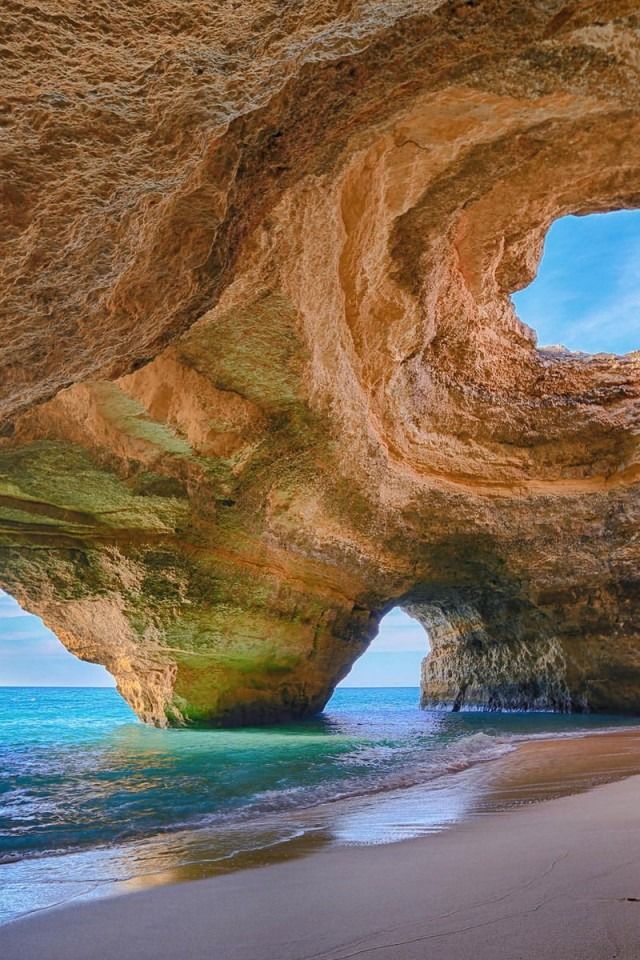 a view from inside a sea cave, looking out to the ocean. the cave is large and has a sandy beach floor. the water is clear and blue, and the sky is a bright blue. the cave is made of orange and brown rock, and there are a few smaller holes and crevices in the rock. the cave is a popular tourist destination in the algarve region of portugal.