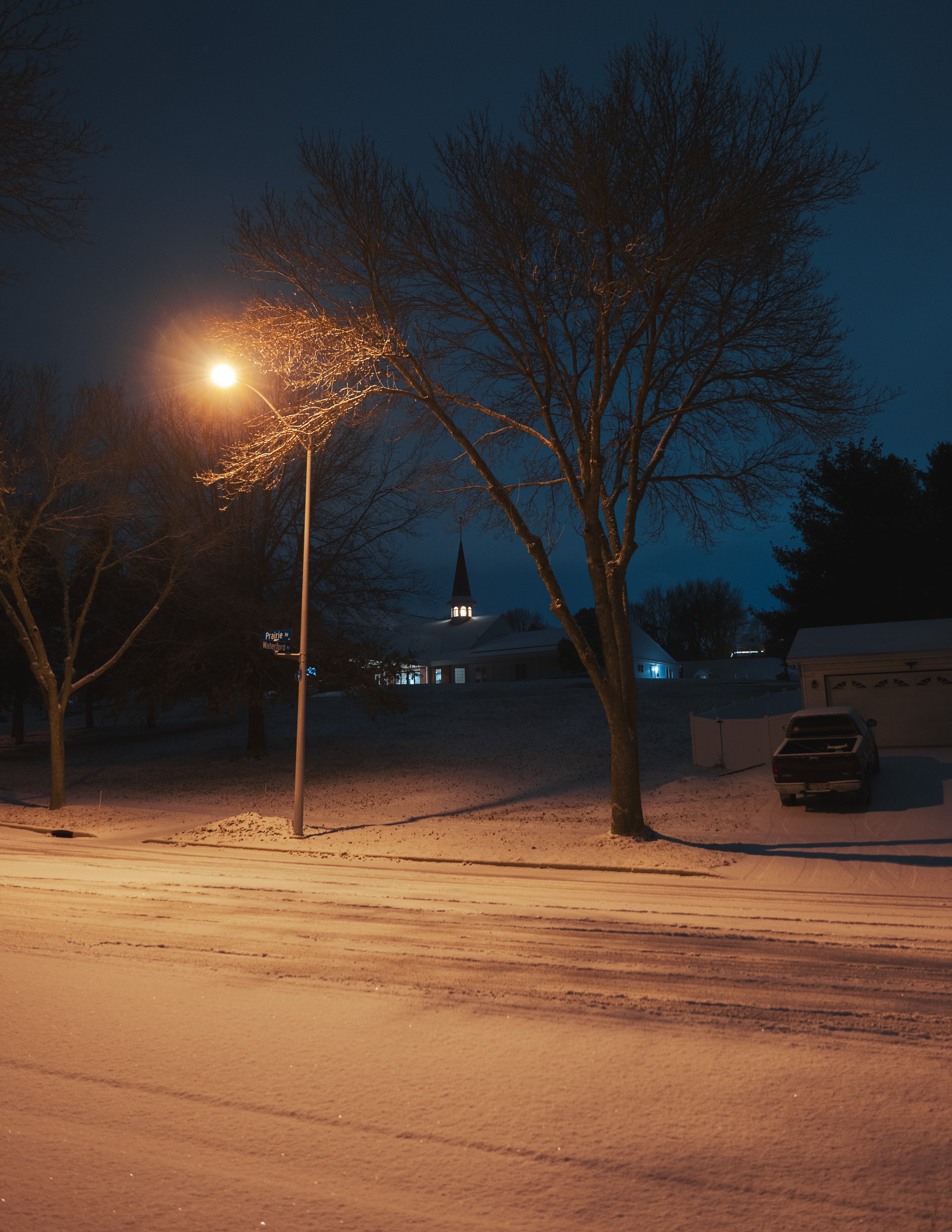 a night scene with a snowy street and a street light illuminating the area. a bare tree stands beside the light, and a car is parked near a building in the background. a church spire is visible in the distance.