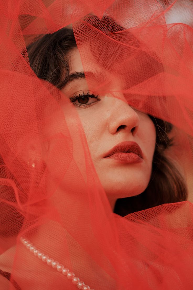 the image is a closeup portrait of a woman's face partially obscured by a sheer, bright red tulle fabric.  the fabric drapes around her head and shoulders, creating a dramatic and somewhat mysterious effect.  her dark brown hair is visible beneath the tulle, and her makeup features dark eyes and bold red lipstick. she wears a pearl necklace visible at the bottom of the image. the overall mood is sensual and artistic, with a focus on the textures of the fabric and the woman's features.  the lighting is soft and diffused, enhancing the dreamy atmosphere.