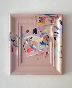 this is a picture of a framed painting with a modern abstract design. the painting features pink, white, blue, red, orange, green, yellow and black brushstrokes. the painting is set against a white wall. the painting is in a pink frame.