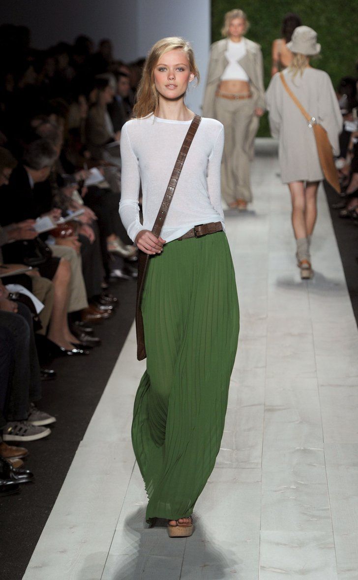 a blonde model walks the runway wearing a white longsleeved shirt, a long green pleated skirt, and a brown shoulder bag. she has blonde hair and is wearing minimal makeup. the background includes other models, and the audience. the setting is a runway show.