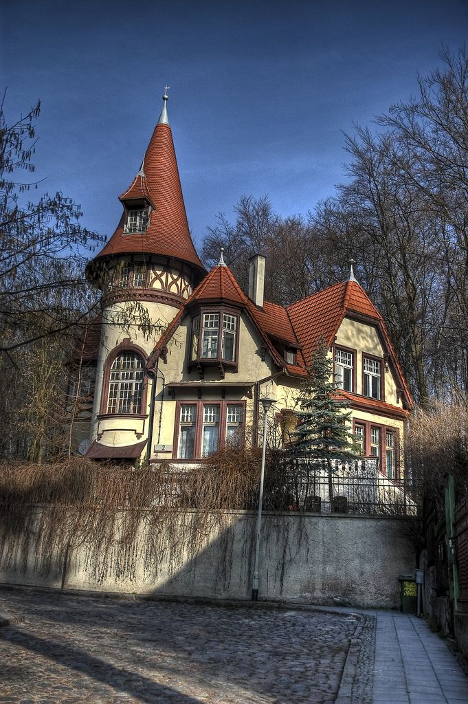 a charming house with a red roof and turret, nestled among trees and bathed in the warm light of a sunny day. the house features intricate details and is surrounded by a lush garden. the street in front is paved with stone, leading the viewer's eye towards the inviting entrance.