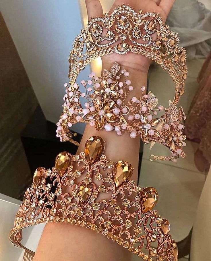 the image showcases three exquisite crowns, each boasting a unique design and color palette, held by a hand.  the top crown is a large, ornate rose gold piece featuring clear and amber rhinestones. below it is a smaller, more delicate rose gold crown with pink accents and beads. at the bottom, another rose gold crown is displayed, this one with larger ambertoned stones. all three crowns are richly detailed, displaying intricate filigree work and sparkling gemstones. the overall aesthetic is glamorous, luxurious, and bridalthemed. the image is a closeup shot, highlighting the intricate details of each crown.