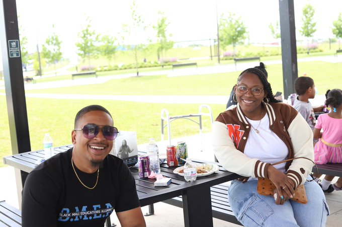a man and a woman sit at a picnic table outdoors on a sunny day. the man is wearing sunglasses and a black shirt, and the woman is wearing a letterman jacket. they are both smiling and appear to be enjoying themselves. there are other people in the background, including children, and there is food and drink on the table. the setting is a park with green grass and trees.