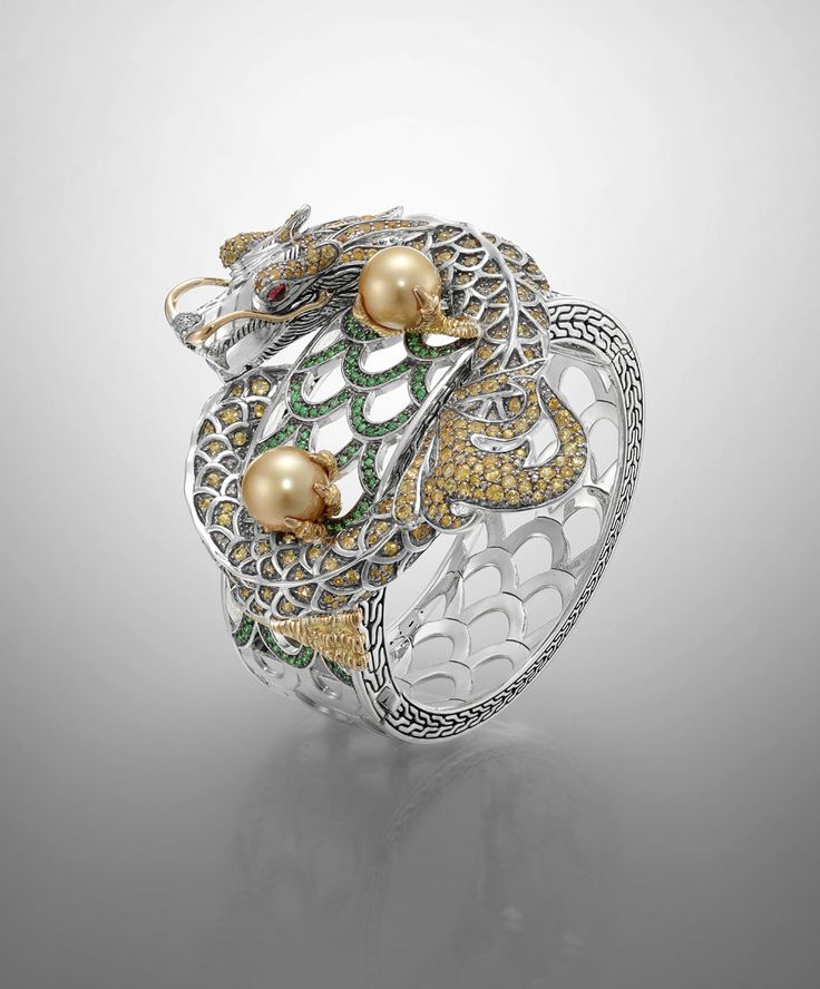 a highly detailed and ornate bracelet featuring a dragon motif. the dragon is crafted in silver with gold accents, and it holds two large golden pearls. the bracelet is embellished with green and yellow gemstones. the design incorporates intricate filigree work and has an openwork pattern on the sides. the overall aesthetic is luxurious and asianinspired.