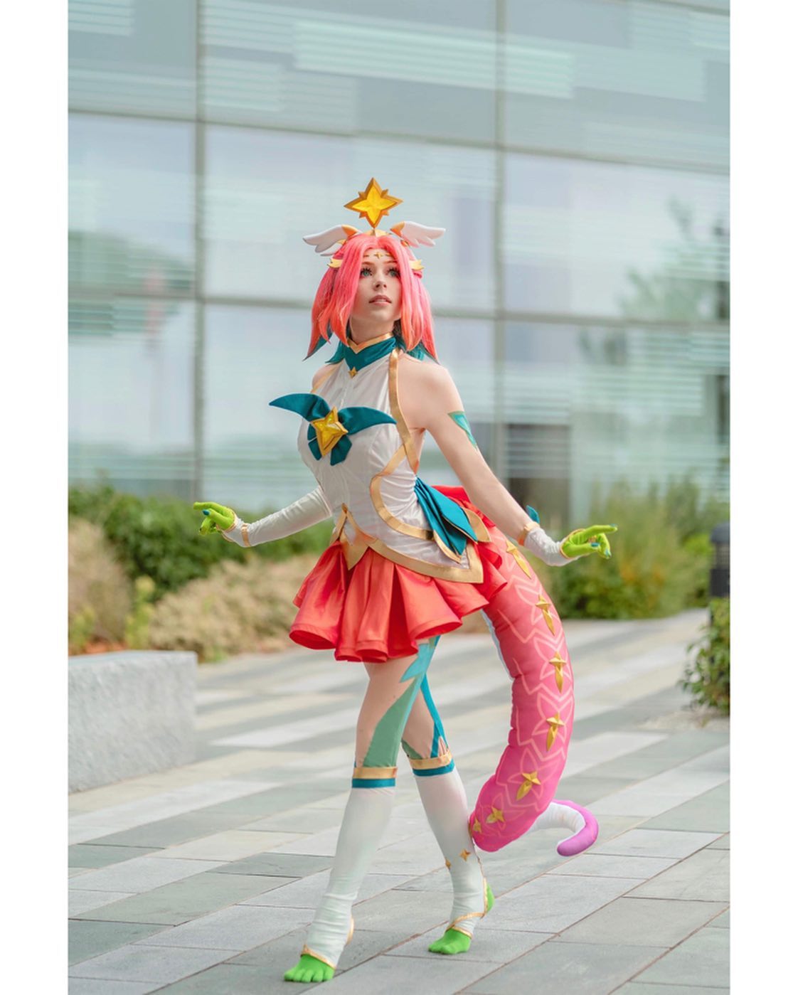 the image shows a female cosplayer dressed as star guardian soraka from the video game league of legends.  the cosplay is highly detailed and accurate, featuring soraka's signature pink and white outfit, star accents,  and a long, pink tail. the cosplayer is posing outdoors in a modern urban setting, against the backdrop of a glass building.  the lighting and setting give the image a clean and professional feel, highlighting the quality of the costume.  the cosplayer's pose and expression convey the whimsical and magical nature of the character.