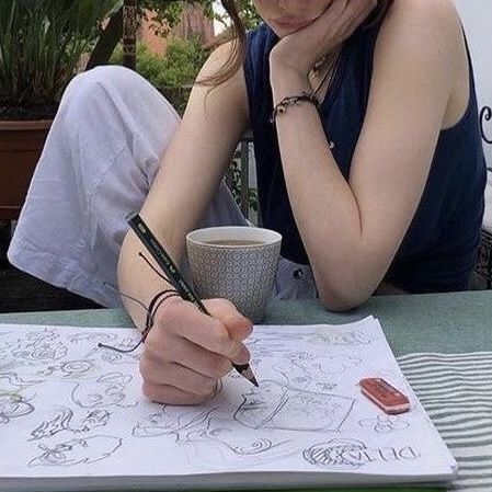 a woman is sketching in a notebook with a pencil outdoors. she has a cup of coffee beside her and is wearing a bracelet. the notebook is full of various sketches, and an eraser sits on the page as well.