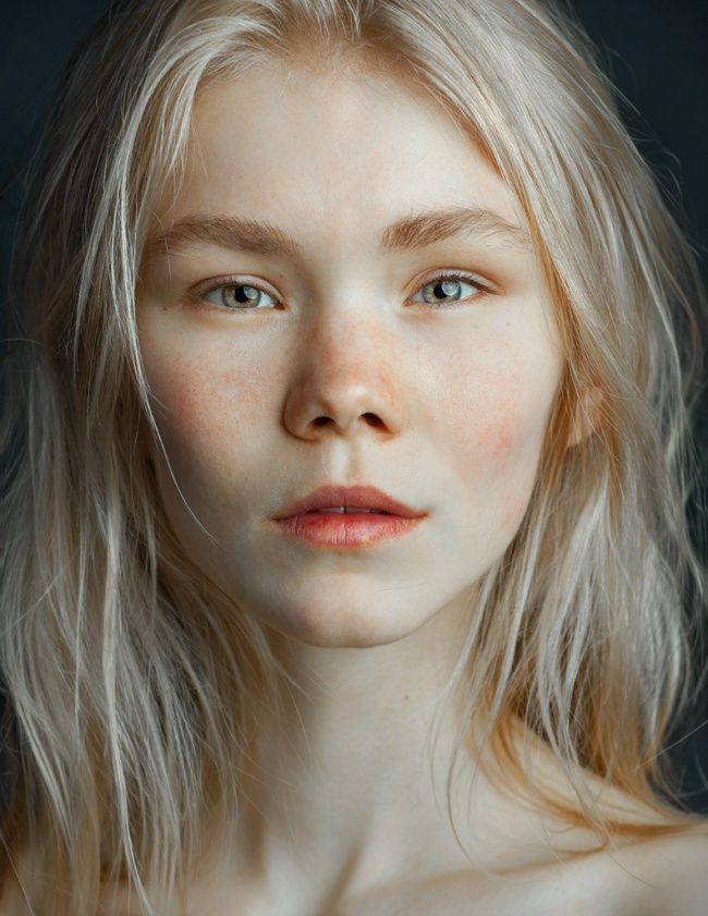 a closeup portrait of a young woman with blonde hair and fair skin. she has freckles and lightcolored eyes. her expression is serious. the shot is a headshot, focusing on her face and upper chest.