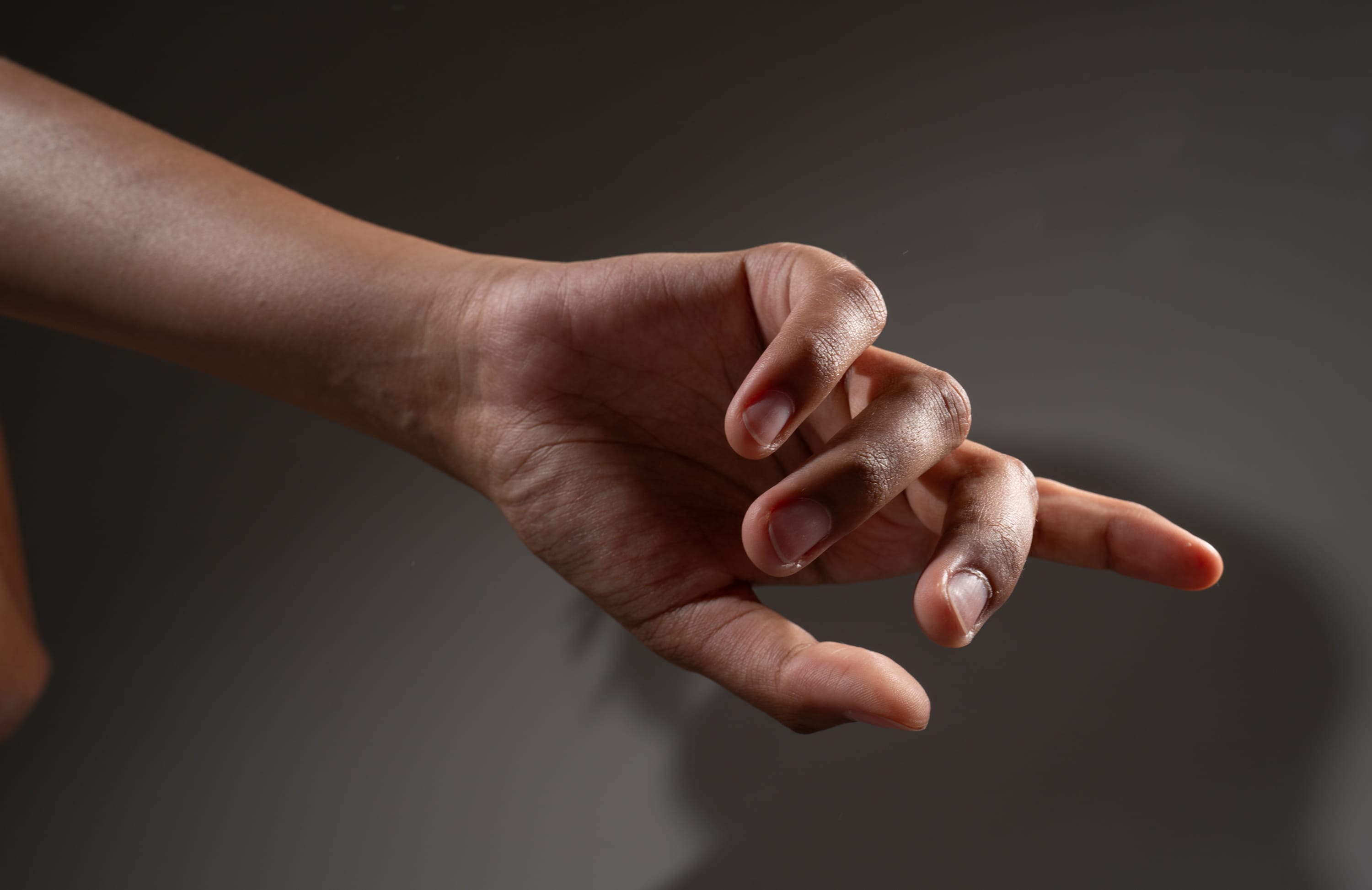 the image shows a hand with dark skin reaching out against a gray background. the hand is illuminated by studio lighting, creating a sense of tension and drama. the fingers are slightly curled, as if the hand is about to grab something.