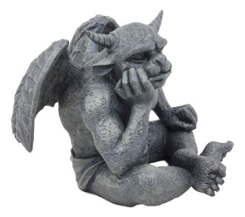 a gray stone gargoyle statue sits with its legs crossed and one hand propping up its chin, as if in a thoughtful or pondering pose. it has wings, horns, and a detailed, textured surface. the statue appears to be a decorative garden or architectural element.
