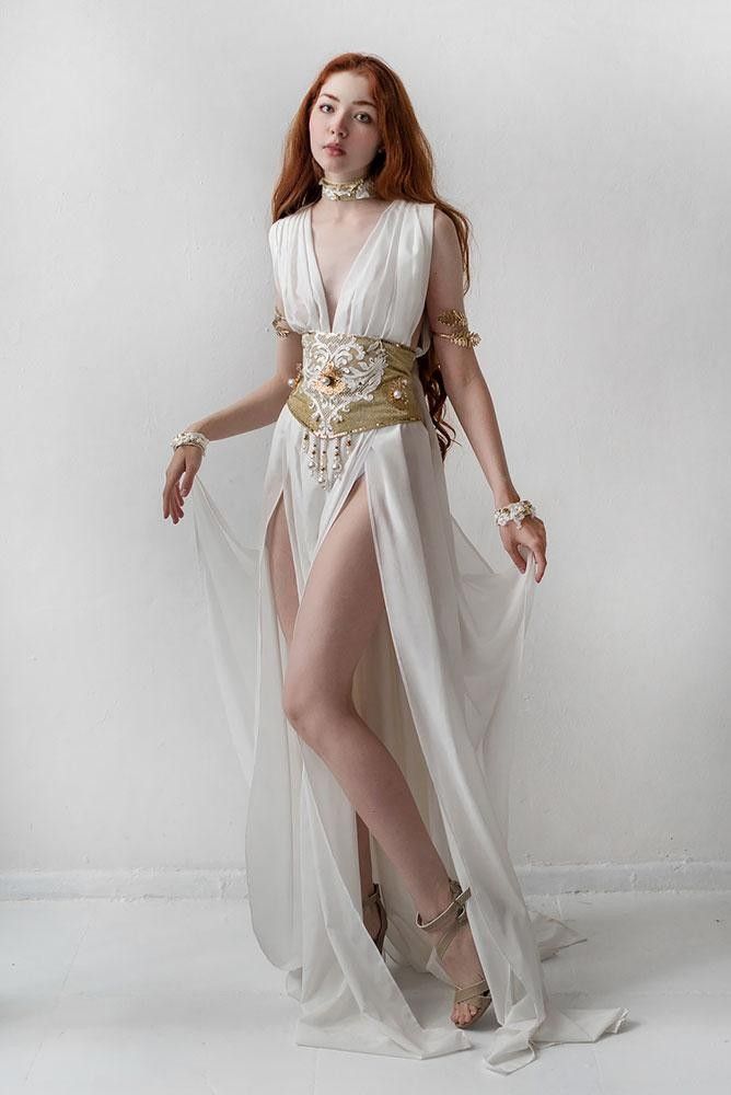 a fulllength shot of a redhaired woman wearing a flowing white dress with a high slit and an ornate gold belt. the dress is reminiscent of an ancient greek or goddessinspired design. she's accessorized with gold bracelets, a choker, and sandals, and her pose is graceful and elegant. the setting is a simple white studio background, making the dress and model the focal points of the image. the overall aesthetic is ethereal and fantasylike.