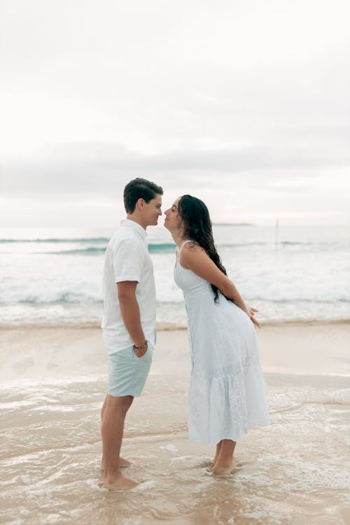a young couple stands in the shallow water of a beach, facing each other. they are about to kiss, and their faces are close together. the man is wearing a white shirt and light blue shorts, and the woman is wearing a flowing white dress. the ocean waves are gently lapping around their feet. the overall scene is serene and romantic.