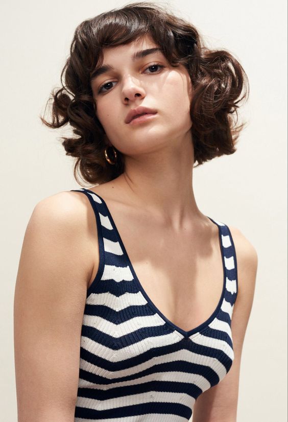 a young woman with short curly brown hair  is posing in a blue and white striped tank top. she is looking directly at the camera. the background is a pale yellow.