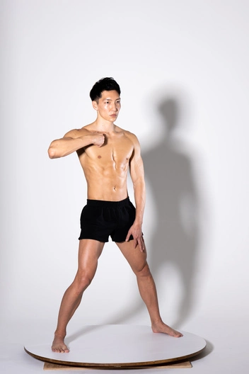 the image shows a young, muscular man posing in a studio against a plain white background. he is shirtless, wearing only black athletic shorts. his physique is welldefined, showing a clear sixpack and overall lean musculature.  he is standing on a round white platform with his weight slightly shifted, suggesting a posed shot, possibly for fitness or modeling purposes. his expression is neutral and confident. the lighting and background create a minimalist aesthetic.