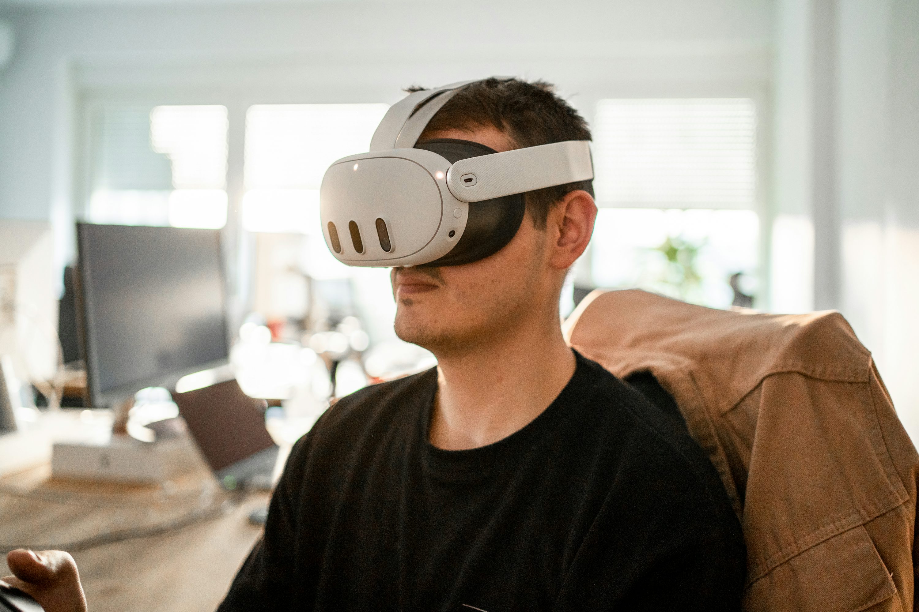 a man is wearing a white virtual reality headset while seated at a desk indoors. the desk has a computer, laptop, and controller on it. he is wearing a black shirt and has a tan jacket draped over his chair. the scene is welllit by natural light coming through a nearby window.
