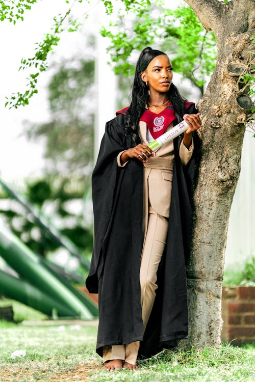 the image shows a young african woman, elegantly dressed in a black graduation gown over a beige pantsuit, standing confidently outdoors. she holds a rolledup diploma or certificate.  the background is blurred but shows a tree trunk she's leaning against, green grass, and some outoffocus structures in the distance. the woman appears proud and happy, celebrating her academic achievement. the logo on her gown suggests it is a university of pretoria graduation.  she is barefoot.