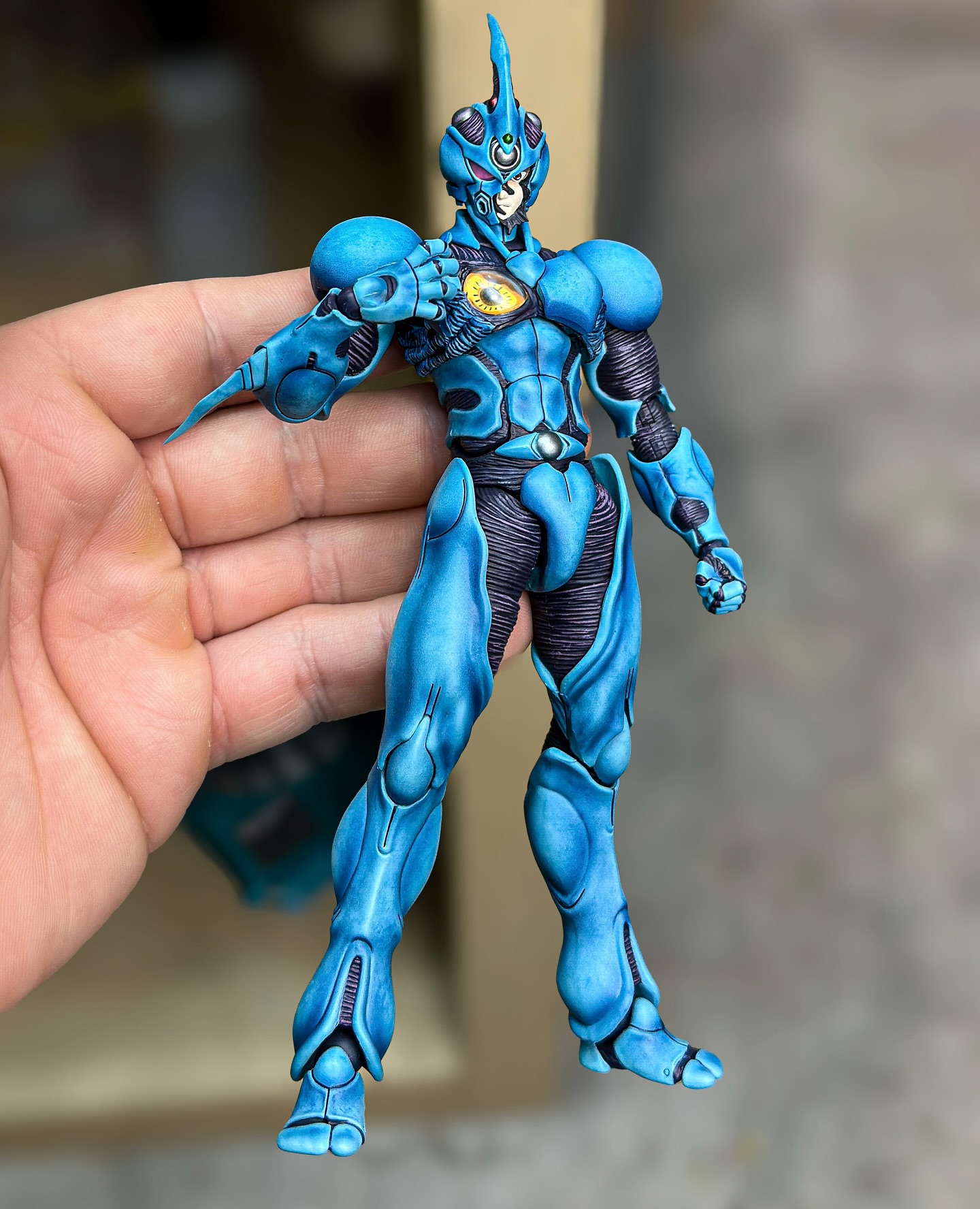 a blue cyborg action figure is being held by a hand. the action figure has a unique design with organiclooking armor, a horn on its head, and detailed mechanical features. the figure is mostly blue with black accents. the hand holding the figure is fairskinned. the background is out of focus.