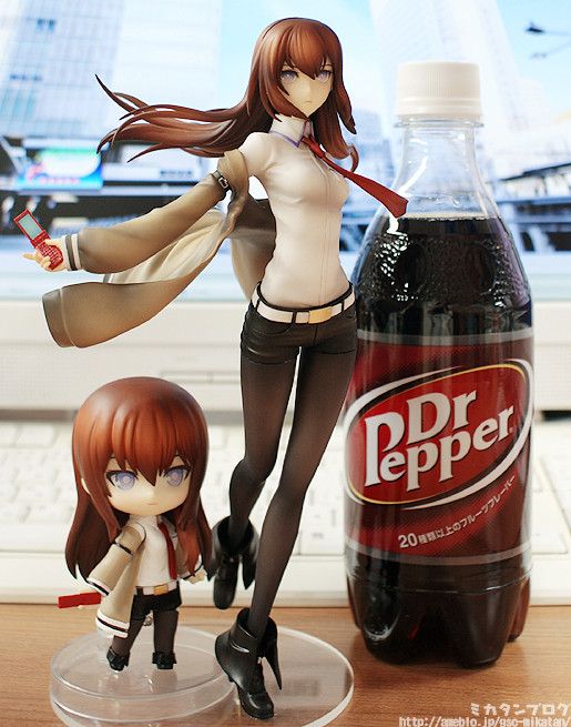 the image shows two figures of kurisu makise from the anime steins;gate.  a larger, more detailed scale figure stands next to a smaller nendoroid version. both figures are posed dynamically.  a bottle of dr. pepper is placed between the figures, providing a scale reference and a contrast in style. the background features a blurry office setting with a computer and keyboard visible. the overall aesthetic is one of careful product photography, showcasing the collectible figures and using the soda bottle to give a sense of scale.