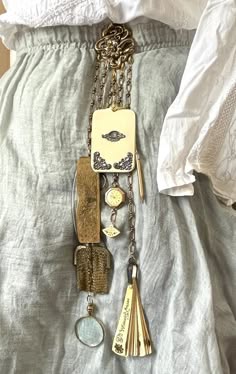 a close up shot of a vintage necklace that hangs on a white blouse. the necklace is made of a series of charms and pendants that includes a pocket watch, a locket, several keys, and a magnifying glass.  the charm are all connected by a delicate gold chain. the blouse is adorned with lace and embroidery. the image is a closeup and shows the detail of the jewelry in great depth.