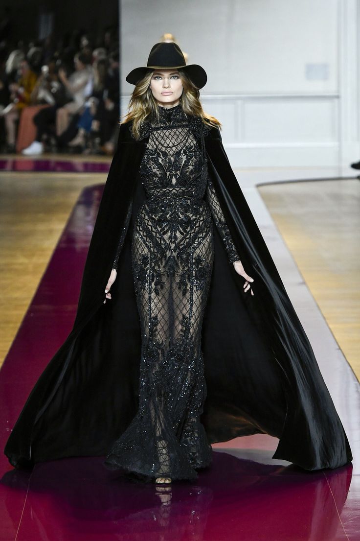 a woman in a long black gown with a black cape walks the runway during a fashion show. she is wearing a black hat and has her hair pulled back. the gown is very intricate with lace and beading throughout. the cape is flowing behind her and adds an elegant touch to the ensemble. the woman is wearing many accessories, including jewelry.