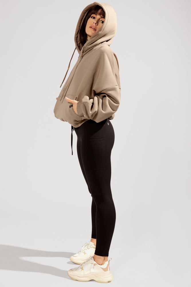 a young woman poses in a beige hoodie and black leggings. she is wearing white sneakers. she is standing in a studio setting.