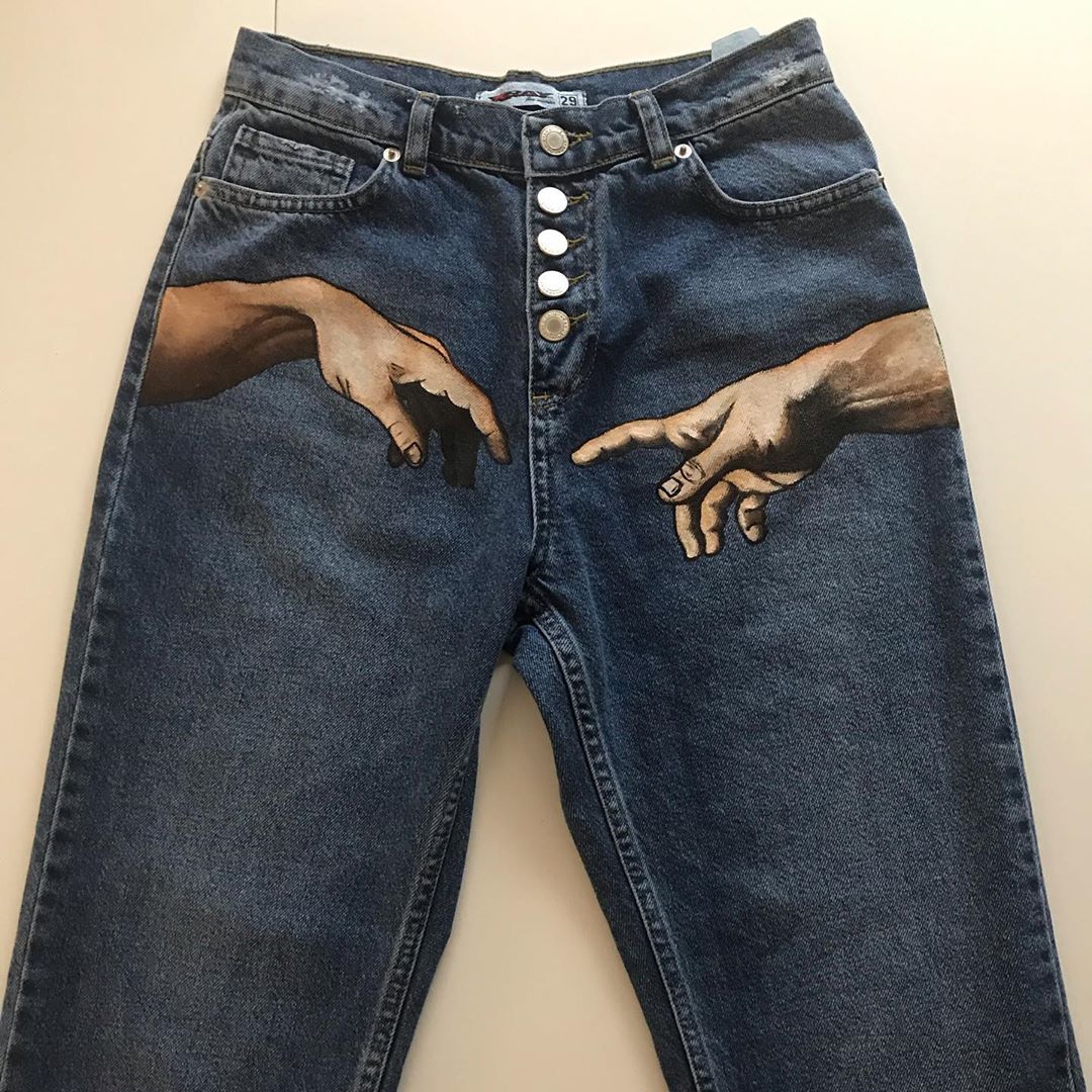 a pair of denim jeans lies flat, showcasing a handpainted rendition of michelangelo's "the creation of adam" on the front. the iconic hands reaching towards each other are centrally placed, adding an artistic flair to the otherwise ordinary jeans.