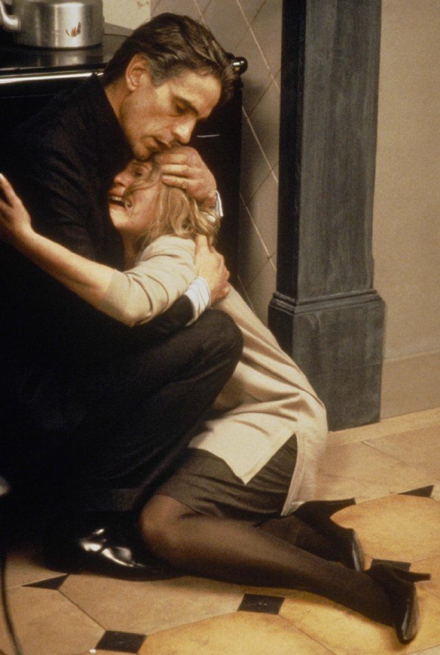 a man in a suit is embracing a woman in a beige coat and black stockings,  their faces are both downcast and the woman appears to be crying.  the man's face is buried in the woman's hair, as she holds him tightly.  they are standing in an indoor setting, with tiled floors and a wooden door behind them.  the lighting is dim and the colors are muted, creating a somber mood.