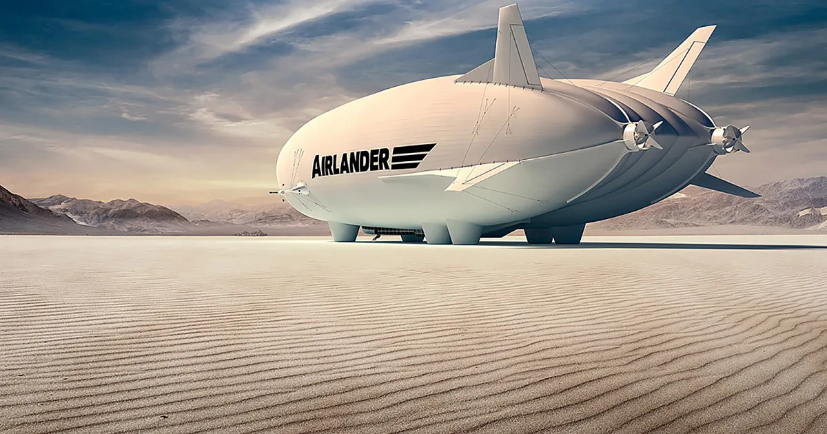 a large, white airship named airlander 10 sits on the ground in a desert setting. the airship is partially deflated, and its engines are visible. the background features mountains and a clear, blue sky with clouds. the sand in the foreground is rippled and textured.