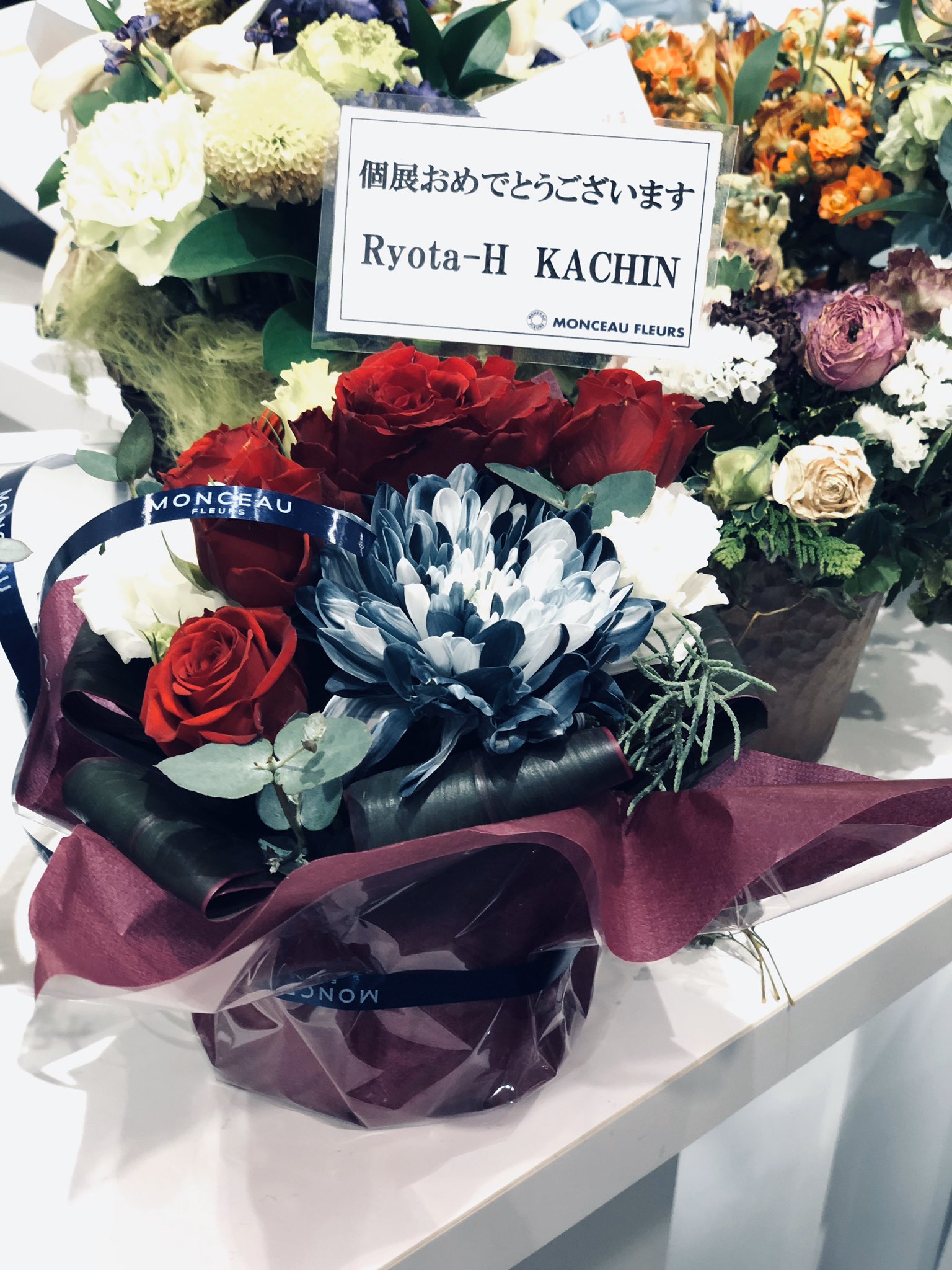 a flower arrangement of red roses and a blue and white chrysanthemum in full bloom, wrapped in dark red wrapping paper and tied with a navy blue ribbon. a white card with japanese text expressing congratulations is attached to the arrangement. the bouquet sits on a white table with other flower arrangements blurred in the background.