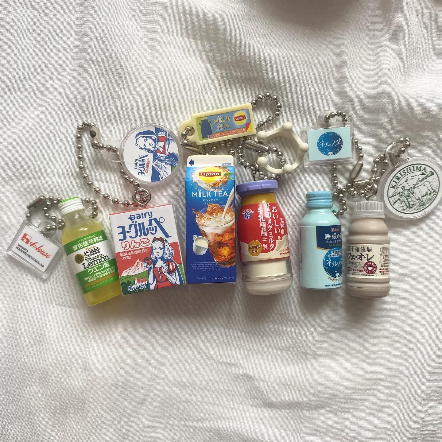 the image shows a collection of keychains, each representing a different japanese dairy or beverage product. there's a miniature carton of lipton milk tea, a box of dairy yogurt, a bottle of c1000 vitamin lemon drink, several other dairy products in small bottles with japanese labeling, and keychains with logos for kirishima and the house brand. all the keychains are laid out on a white cloth surface.