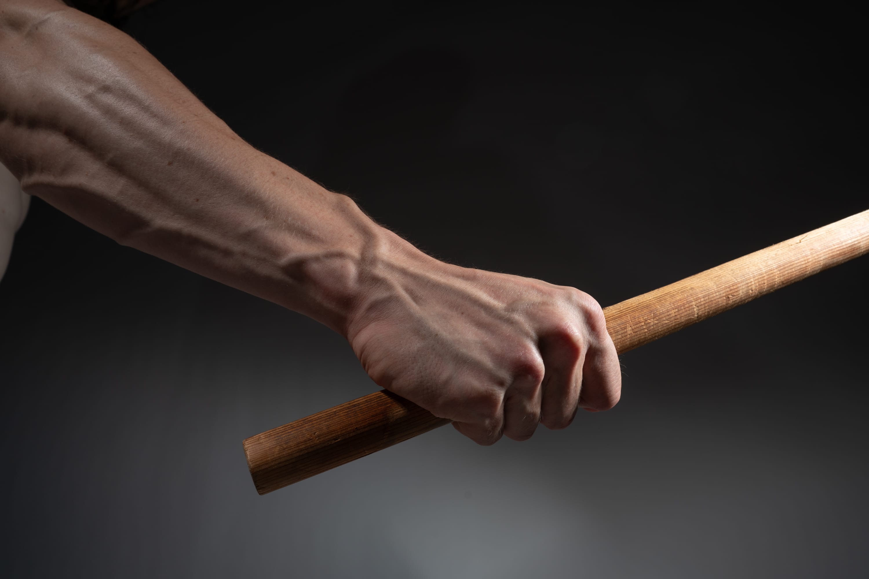 a closeup shot of a muscular man's arm and hand tightly gripping a wooden stick against a dark gray background. the image emphasizes the strength and tension in the hand and arm, conveying a sense of power and potential action.