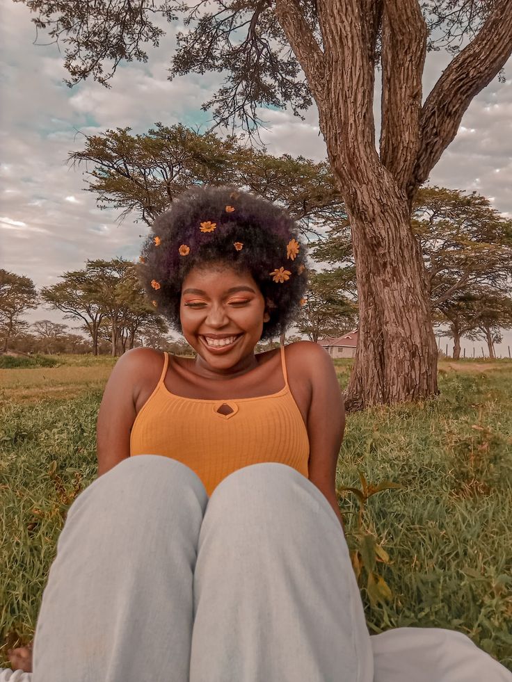 a young woman with a big afro adorned with yellow flowers is sitting in a grassy field, leaning against a tree. she's wearing a yellow tank top and light blue jeans. she's looking down and laughing, with her eyes closed, giving a sense of genuine happiness and joy. the image exudes a warm, natural feel with soft, diffused sunlight.
