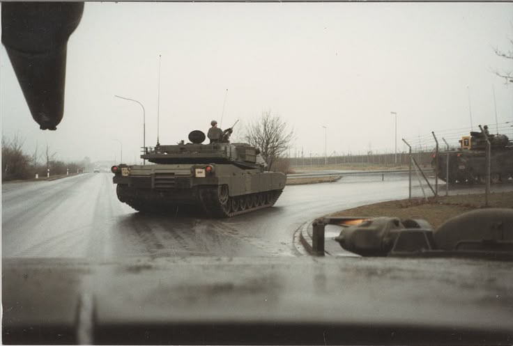 the image shows an m1 abrams tank driving on a wet road in what appears to be germany. it's a rainy or overcast day. another military vehicle is visible in the background.  the perspective is from inside another military vehicle, as part of the front of another vehicle is seen at the bottom of the image. a soldier is visible in the turret of the m1 abrams tank. the overall scene suggests a military convoy or movement.