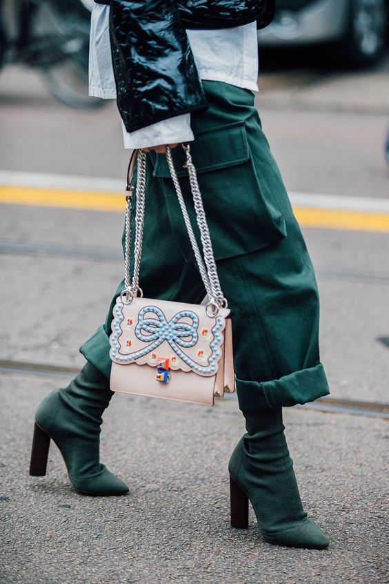 a stylish woman is walking down the street. she is wearing a green outfit that consists of cargo pants cuffed at the ankle and green sock boots with high heels. she carries a light pink fendi bag with a blue bow on it. the overall image is wellcomposed and shows the woman's fashionable look.