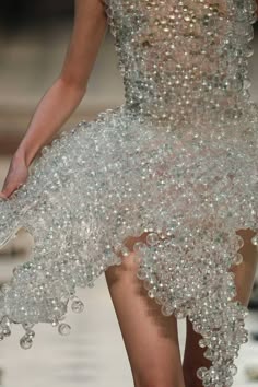 a woman wearing a stunning, beaded dress. the dress is made entirely of small, iridescent beads that create a shimmering, ethereal effect. the model's arm is outstretched, highlighting the texture of the dress. the dress is a work of art and would be perfect for a formal event, like a wedding or a gala.