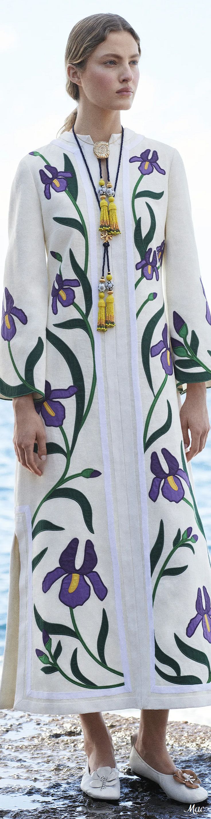 a full shot of a woman wearing a long white dress with embroidered irises. the dress has long sleeves and a relaxed fit. the woman is standing outdoors near the water, and she is wearing simple shoes and a long necklace with tassels.  her hair is pulled back in a low ponytail.  the overall style is elegant and summery.