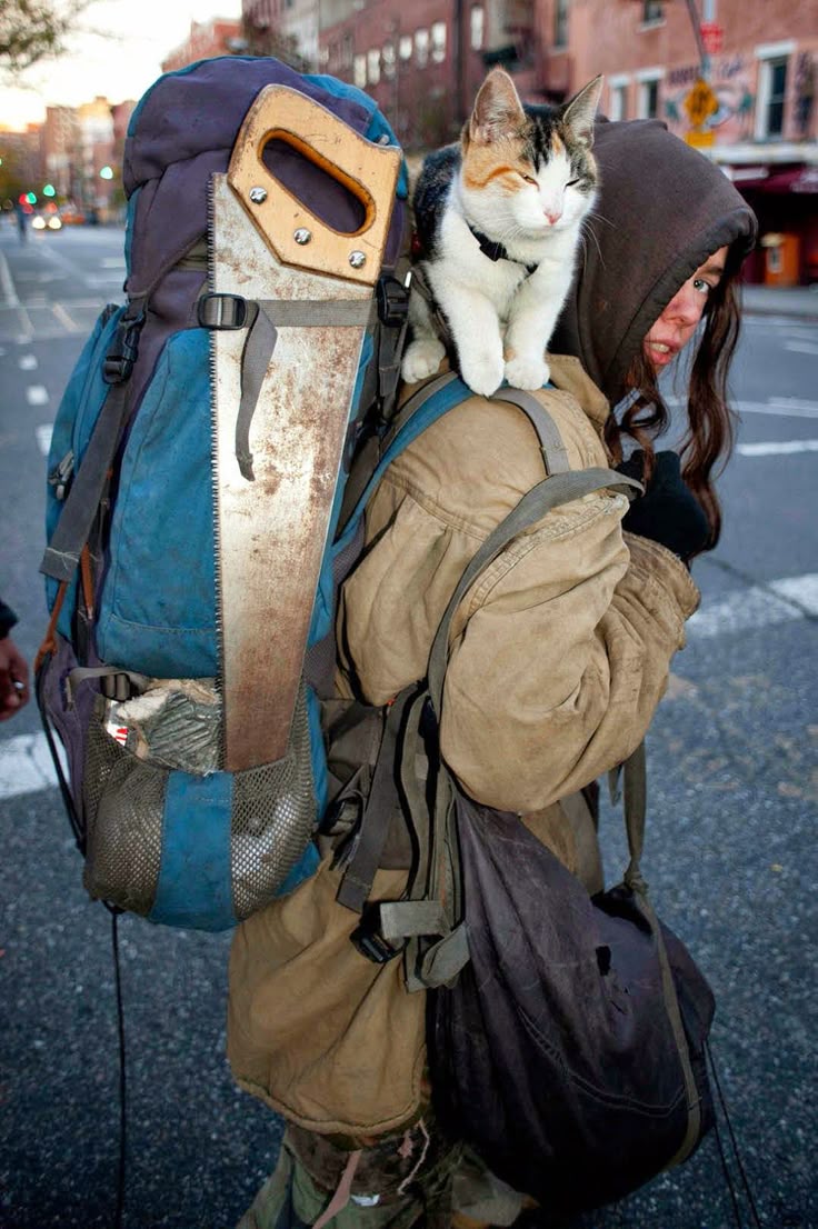 a woman is walking down the street with a large backpack that has a saw attached to it, and a cat sitting on her shoulder. she is wearing a hoodie and gloves, and carrying a bag. the background is a city street.