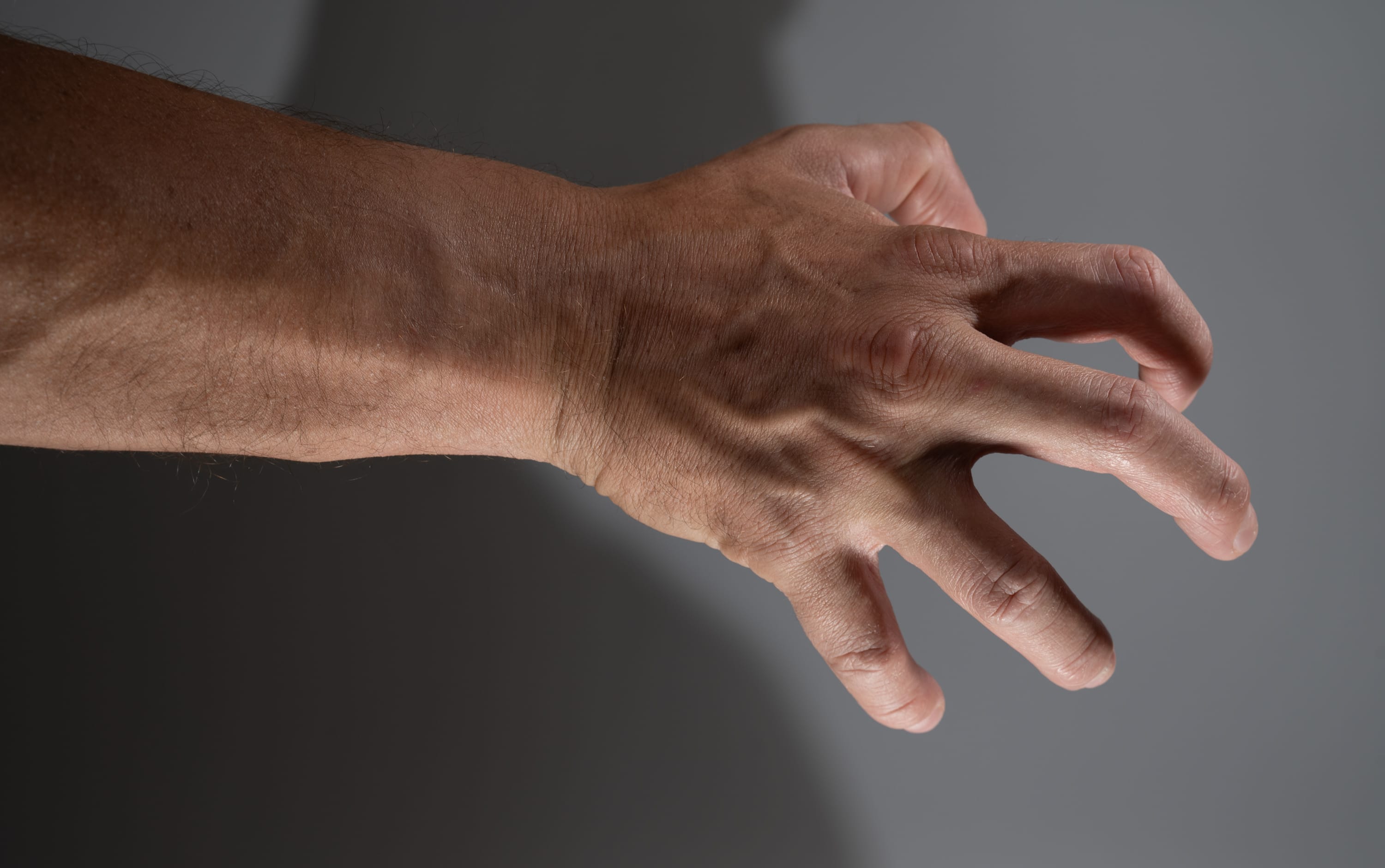a man's left hand and forearm are extended against a gray background. the hand is partially clenched, with the index finger and thumb touching and the other fingers curled inward. the veins on the hand and arm are prominent. the lighting is from the left side, casting a shadow on the right side of the image.