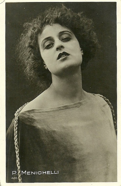 a black and white vintage portrait of a young woman in a studio setting. she is looking away from the camera, with her head tilted back. she has dark, curly hair, and is wearing a dress with a beaded necklace.