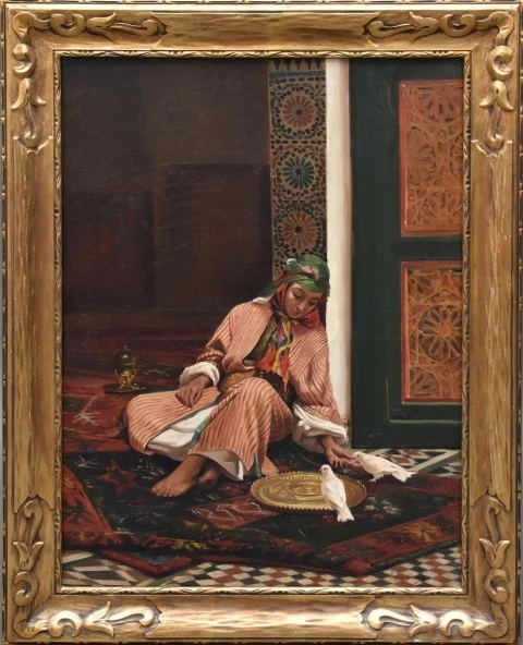 the image is a framed painting of an orientalist scene. a woman in traditional clothing is seated on a carpet in an interior space. she is surrounded by doves, and there is an ornate brass tray nearby. the background features a doorway with intricate patterns and a gold frame surrounds the painting.