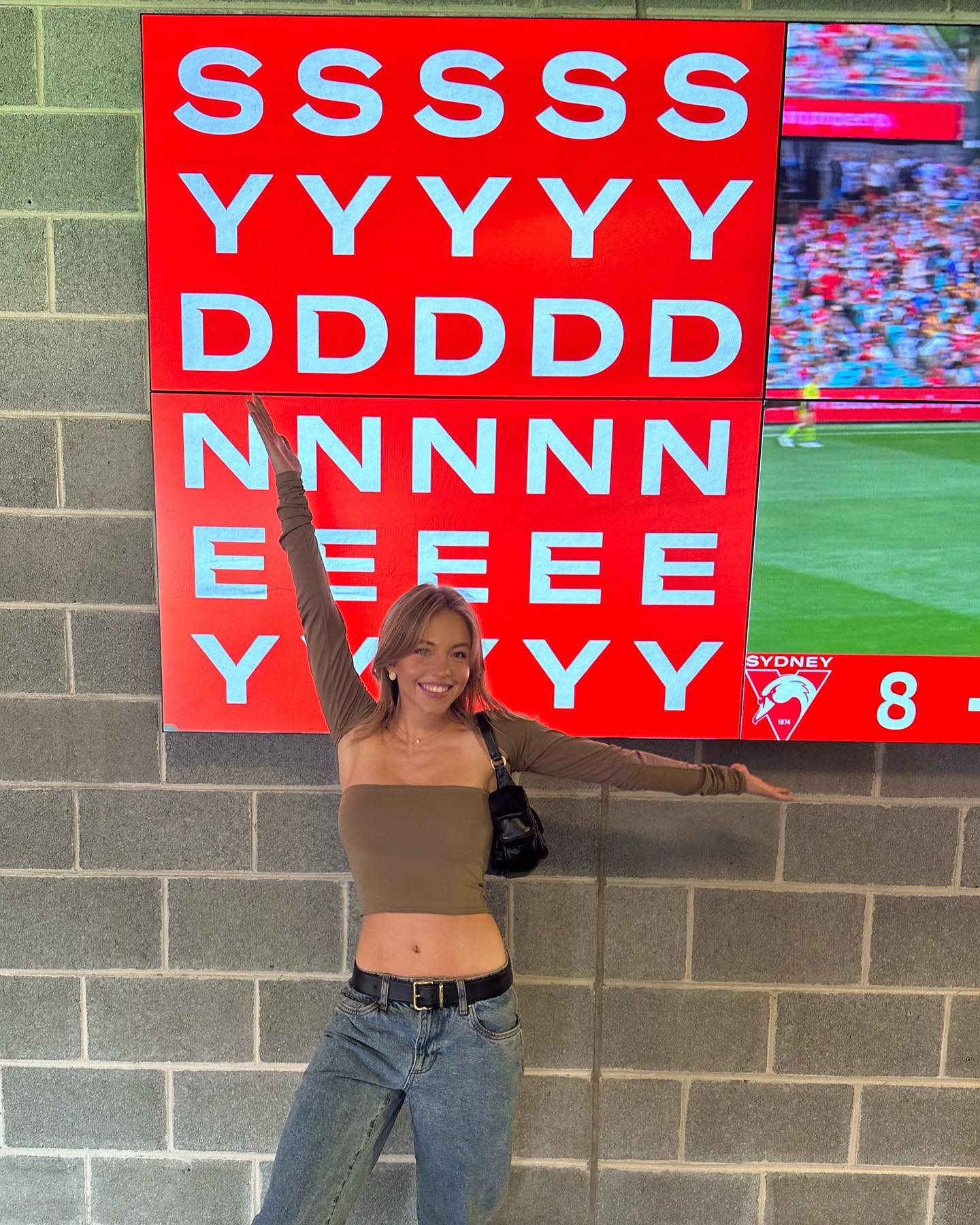 a young woman in a stylish outfit poses in front of a large red sign displaying repeating letters. the sign is located on a brick wall, and behind it are screens showing a soccer/football game. the woman is wearing a brown crop top, baggy light wash jeans, a black purse, and a longsleeved shirt. she is smiling at the camera. the overall atmosphere is casual and trendy.