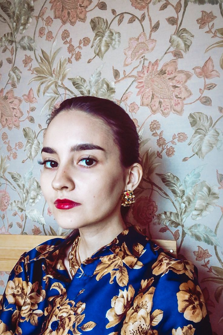 closeup portrait of a young woman with dark hair pulled back, wearing a vibrant blue floral shirt, gold earrings, and a gold necklace. she has red lipstick and a serious expression. the background is a vintage floral wallpaper with a muted color palette. the image is welllit and sharply focused, highlighting the details of her clothing and makeup. the overall mood is one of sophisticated style and quiet confidence.